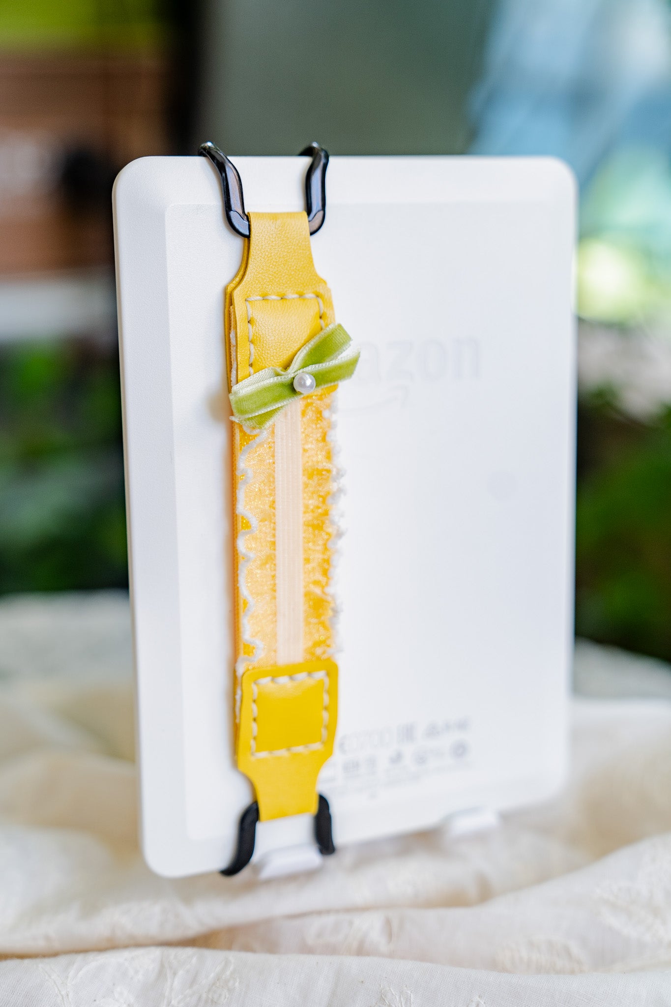 Closeup detail of Kittentailor handmade Lemon Kindle strap showing durable metal clips, soft yellow leather with matching double-layer elastic, green bow, pearl, and lace.