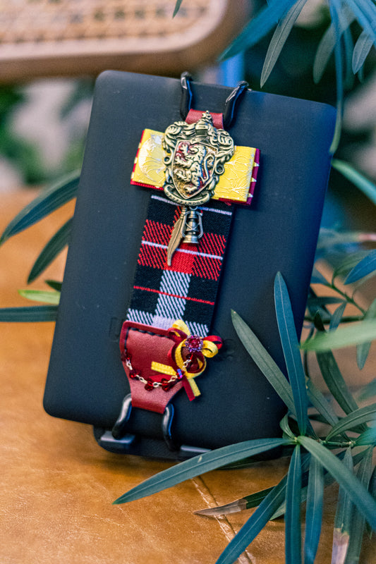 Kittentailor vintage plaid Kindle strap featuring red and black tartan, yellow ribbon bow, and ruby charm. Detailed Dark Academia aesthetic accessory with academy badge.